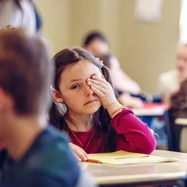 Ein junges Mädchen im Alter von 6 bis 9 Jahren sitzt in einer Schulklasse an einem Tisch und wirkt überfordert und betrübt, während sie sich den Kopf hält. Im Hintergrund sind verschwommene Silhouetten einer Lehrerin und Mitschüler zu sehen. Das Bild zeig